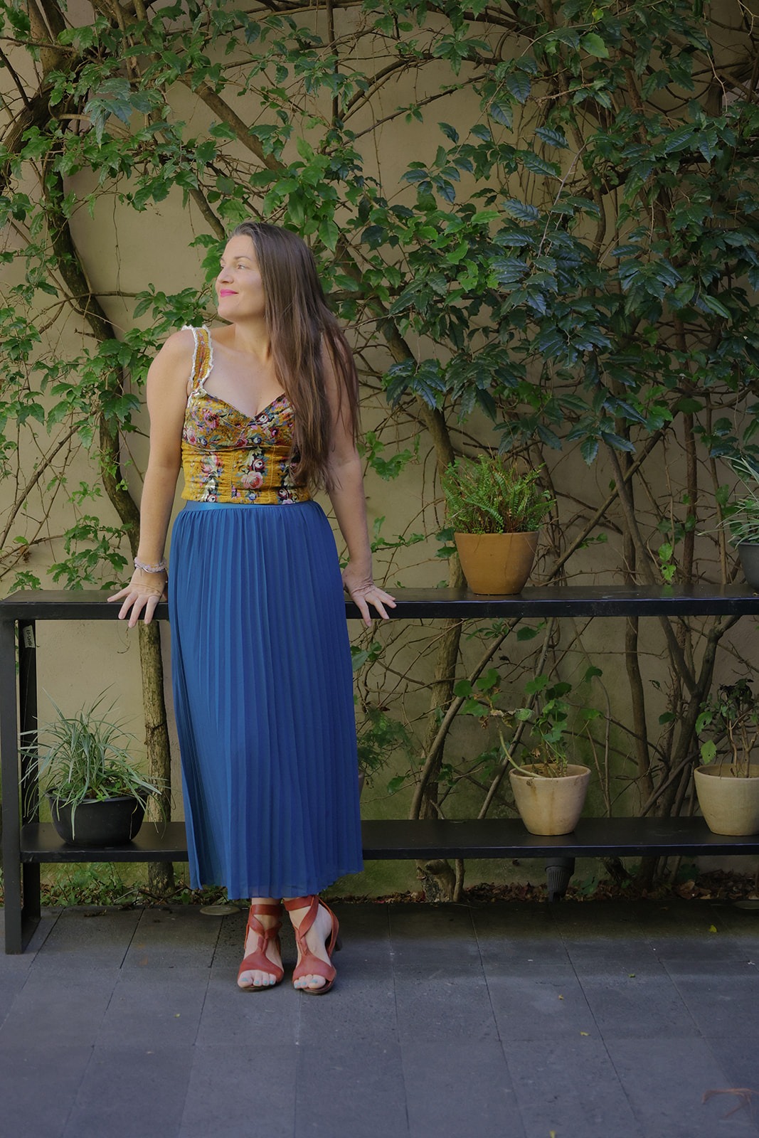 Megan Swan standing at a vine-covered wall in a blue pleated skirt